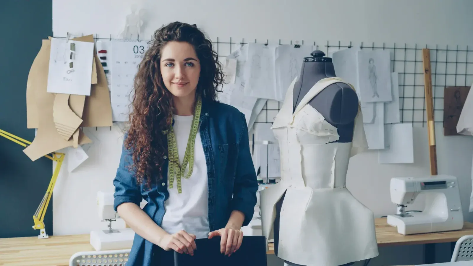 A tailor stands in a workshop beside a half-draped dress form, a measuring tape around her neck, with pattern sketches and sewing machines behind her